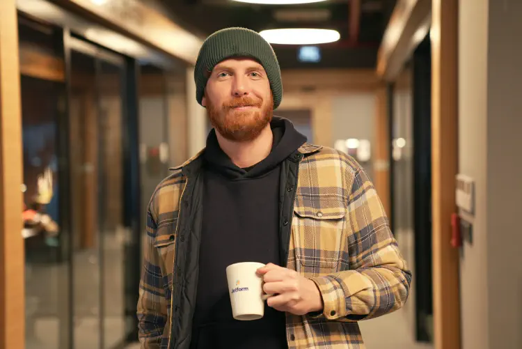 Picture of a white male in a hat and flannel jacket smiling. He has a red beard and is holding a coffee cup that says jotform on it. He could be a small business owner or manager.